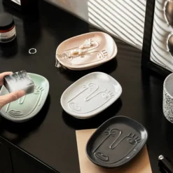 Collection of four Motessa abstract face ceramic trays in blush pink, sage green, ivory white, and charcoal grey, styled with soap and jewelry.