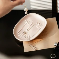 Motessa blush pink ceramic abstract face catchall tray beautifully displaying a silver necklace and pearl earrings in warm sunlight.