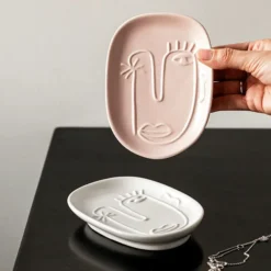 A hand holding up a Motessa blush pink abstract face tray above an ivory white ceramic tray on a dark surface.