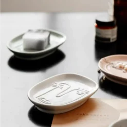 Motessa ivory white and sage green abstract face trays styled as a soap dish and jewelry holder on a dark vanity counter.