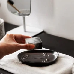 A hand placing a marbled soap bar onto a Motessa charcoal grey abstract face ceramic soap dish next to a modern vanity mirror.