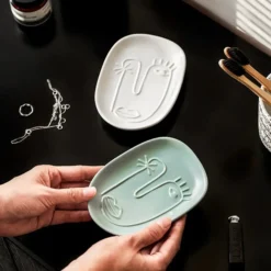 Hands holding a Motessa sage green abstract face tray in a minimalist bathroom setting with a white tray and bamboo toothbrushes.